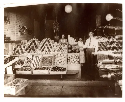 Man and two women standing in the midst of crates of fruit and vegetables placed at the open front of a shop. Scales are visible for weighing.