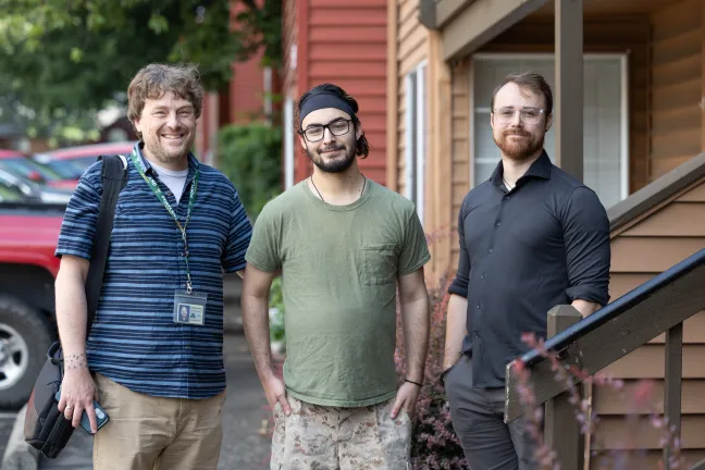 A medium outdoor shot shows three men standing side-by-side on a sidewalk in front of a house.