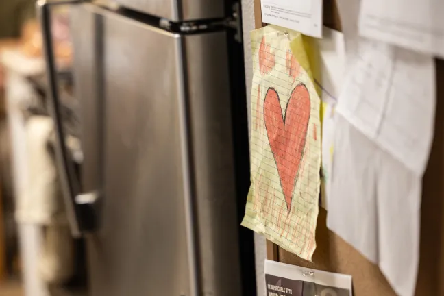 A closeup, eye-level shot of a cork bulletin board, with a large, drawing of a red heart on yellow graph paper pinned to it.