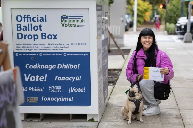 A woman crouches next to a ballot drop box. She holds a yellow envelope, and a small dog sits calmly beside her.