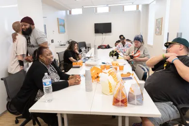 Several people sit around a white table with food from Popeyes; on the left, two people hug to greet each other