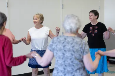A group of five people, dressed in casual clothing and skirts, holds hands in a circle as they participate in a folk dance class indoors