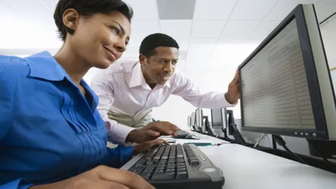 a woman sits at computer while a man stands next to her smiling