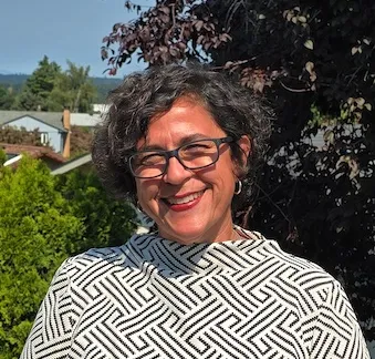 Amara, a light brown skinned middle aged woman with short brown curly hair smiles outside. She is wearing a white shirt with geometric designs and black rimmed glasses.